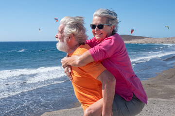 Happy elderly couple at the seaside, husband takes his wife on his shoulders and both  smile and...