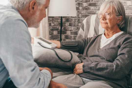 Senior woman using medical device to measure blood pressure suffering of hypertension - elderly couple sitting at home on sofa takes care of health