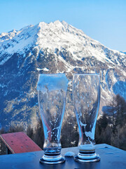Two glasses of beer on a table with a mountain in the background. The mountain is covered in snow...