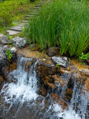 A close-up of a small artificial waterfall in a garden landscape, with water cascading over stone tiers, surrounded by vibrant green grass and lush plants. Stone steps are in the background.