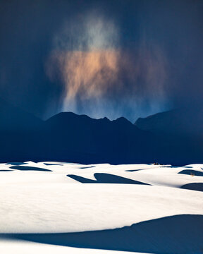White rolling sand dunes and dark mountains in the distance under a rain cloud that has taken the form of a man's face at White Sands National Park, New Mexico. A group of hikers are in the distance.