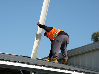 Workman on roof holds metal roofing piece while bending  down to pick up tool.