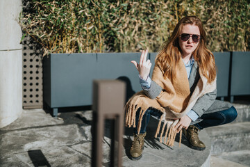 A stylish woman wearing a scarf and sunglasses poses casually in a sunny city environment,...