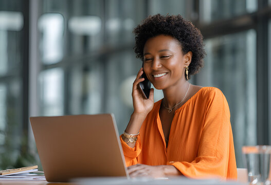 Woman with short hair smiles while using a laptop and talking on a cell phone