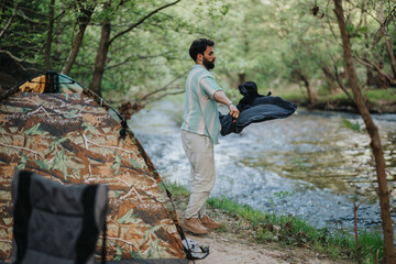 A man prepares a camping tent on a peaceful riverbank surrounded by lush greenery. The scene...