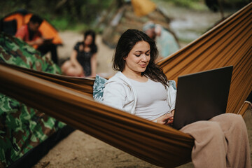 Young woman uses her laptop while lying comfortably in a hammock at a campsite. The setting conveys a balance of work and leisure in an outdoor natural environment, showcasing modern work lifestyle. © qunica.com