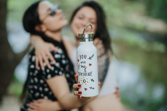 Two individuals share a close moment outdoors, showcasing a creatively designed water bottle emphasizing joy and positivity.