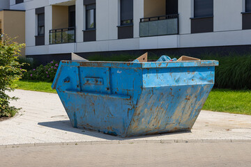 Large rusty blue dumpster container placed near modern apartment building on sunny day. Residential...