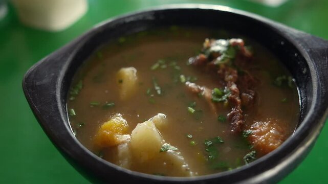 Close-up of hearty sopa de chivo in a rustic black bowl, showcasing vibrant herbs and tender goat meat. Natural lighting enhances the rich texture and warm, inviting atmosphere.