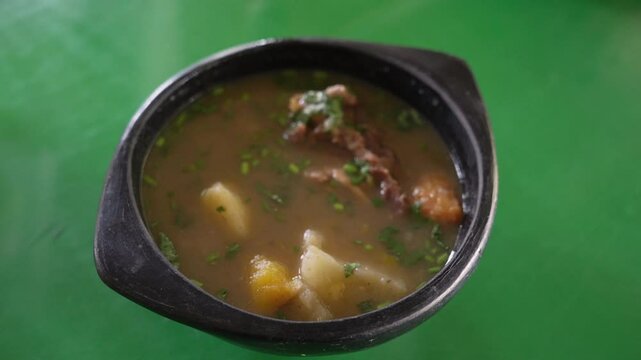 Close-up of steaming sopa de chivo in a rustic bowl. The vibrant dish features rich broth, tender meat, hearty potatoes, enhanced by bright natural lighting on a green backdrop.