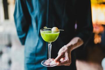 Close-up of a hand holding a cocktail glass with a green drink, garnished with a pickled onion on a skewer. Dark background, moody lighting, elegant presentation.