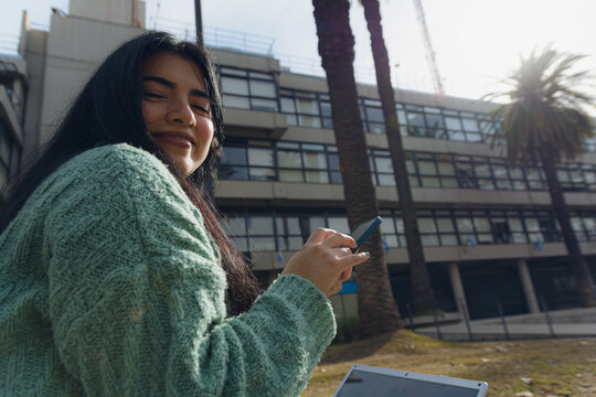 Young woman sitting outdoors using her phone and turning to look at the camera