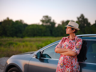 Traveler woman in red ethnic dress and hat drives car through scenic countryside of Croatia in summer. Surrounded by lush fields and forests, she enjoys peaceful and adventurous road trip