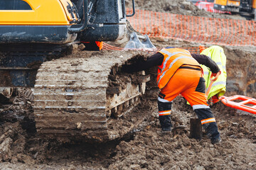 Construction worker  in bright safety gear working hard using shovel to clean tracks of his heavy...
