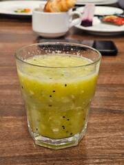 A glass of green juice smoothie kiwi with a few black seeds in it. The glass is on a wooden table