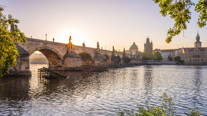 Early morning sun casts a golden glow on the historic Charles Bridge in Prague, reflecting...