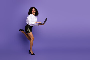 Fototapeta premium Joyful young businesswoman in stylish formalwear jumping with a laptop against a purple background, promoting success