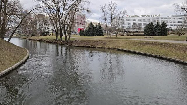 MINSK - MAR 27, 2025: A man is rowing a boat on a river. The river is surrounded by trees and buildings. Sign - Minsk - hero town.