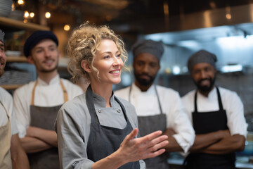 female head chef gesturing while briefing diverse kitchen staff, lively team atmosphere