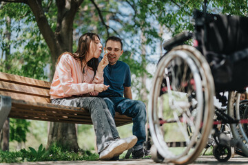 A cheerful couple enjoys a sunny day outdoors sitting together on a bench in the park. A wheelchair...