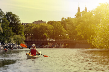 Woman kayaking through a sunlit waterway in Stockholm, with lush trees, moored boats and a...