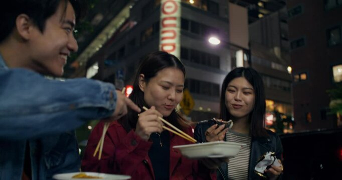Three young people are eating street food and talking while standing in the night city. They laugh and look happy. Tokyo, Japan