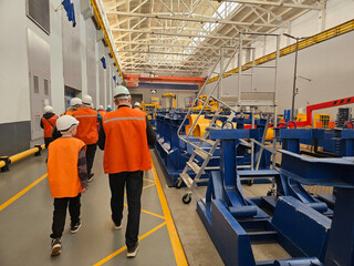 A group of people wearing orange vests walk through a factory. Scene is serious and focused, as the...