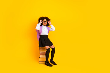 Adorable schoolgirl in stylish uniform posing with glasses and books against yellow background,...