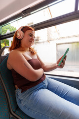 Redheaded woman listening to music, smiling while using smartphone near bus window, relaxed during commute