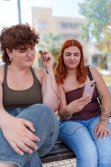Young women sharing earphones while sitting at bus stop, listening to music on smartphone