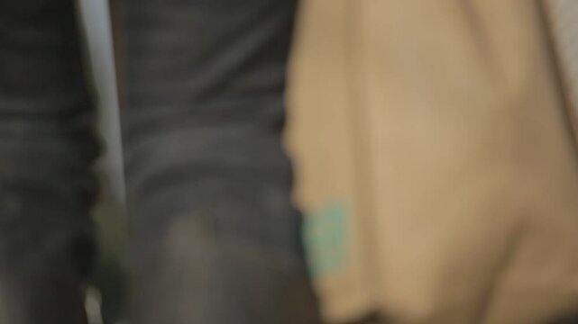 A person, viewed from behind, ascends an outdoor staircase with a brown leather bag slung over their shoulder and several shopping bags in hand. They are dressed in dark jeans and a jacket, with what 