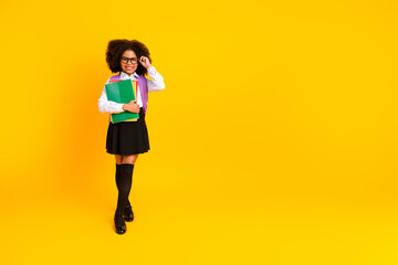 Cheerful schoolgirl in stylish uniform with book and a backpack posing against a yellow background,...