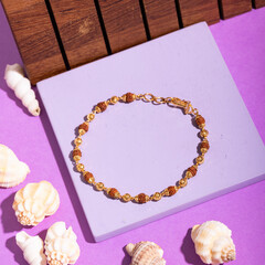 Traditional Rudraksha and gold beaded bracelet displayed on a pastel purple surface, surrounded by seashells, representing spirituality and style.