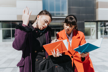 Two young professional women wearing stylish coats, discussing work-related topics with notebooks...