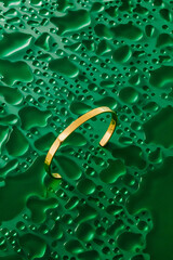 A delicate gold bracelet lies on a vibrant green textured background. The surface features droplets...