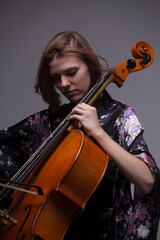 Woman playing cello, wearing floral kimono, against gray background.
