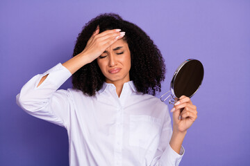 Upset woman with curly hair holding a mirror against a violet background expressing tension in a white formal shirt
