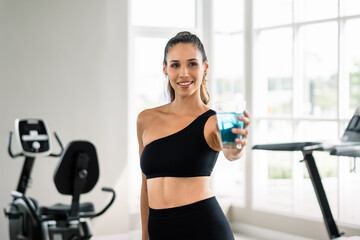 Fototapeta premium A woman is holding a glass of water in a gym. She is smiling and posing for the camera