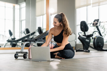 A woman is sitting on a mat with a laptop in front of her. She has a water bottle and a dumbbell...