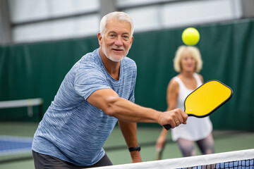 Senior man prepares to hit pickleball with paddle, smiling at partner engaged in game. Indoor facility with green walls, bright lighting, and courts. Concept of sports, fitness, recreation