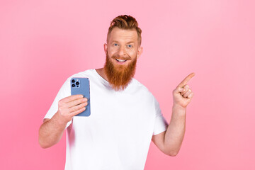 Cheerful red haired man holding a phone in one hand and pointing excitedly with the other against a...