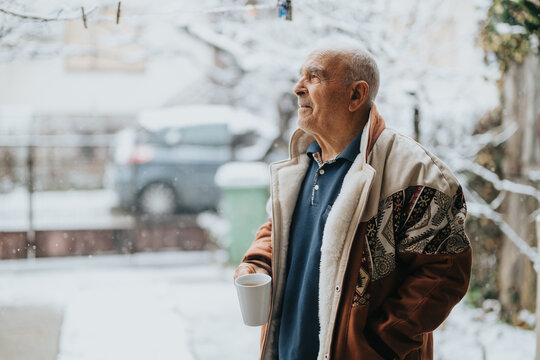 Senior man wearing a patterned coat stands outdoors during snowfall, savoring a warm beverage with a thoughtful expression and serene winter backdrop.