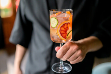 Hand holding a glass of refreshing cocktail with blood orange, cucumber slices, and ice cubes,...