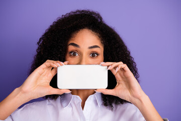 Naklejka premium Confident young woman holding a smartphone in front of her face against a purple background, expressing happiness