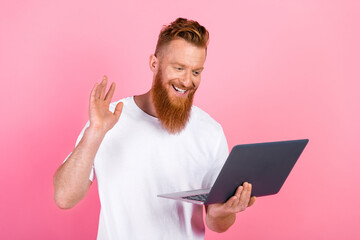 Young red haired man with a laptop on pink background engages in a cheerful video call and waves in...