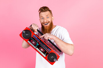 Young red-haired man with beard holding vintage red boombox, smiling joyfully on a pink background...