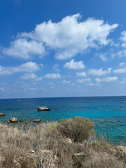 A small boat floats peacefully on turquoise Mediterranean waters under a bright blue sky with scattered clouds, near a rocky, dry coastline.