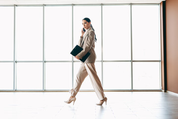 Elegant young businesswoman walking in office while talking on phone and holding folder with...