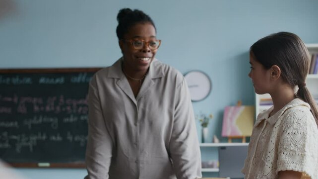 Medium shot of young African American female teacher engaging with Asian schoolgirl unpacking her backpack and getting ready to begin school day
