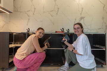 Two women smiling during DIY kitchen cabinet assembly, holding cordless drills, modern interior with marble wall, empowering home improvement, teamwork concept, female builders, real renovation moment